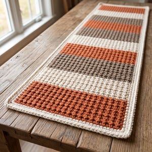 Peut inclure: Un chemin de table rectangulaire en crochet avec un motif en point gaufré texturé. Le design se compose de rayures horizontales alternées dans des tons unis orange brûlé, crème et marron foncé, avec une bordure de couleur crème.