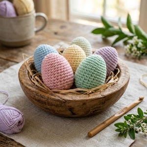 May include: A wooden bowl filled with seven colorful, crocheted Easter eggs. The eggs are pastel shades of pink, blue, yellow, green, and purple. A crochet hook and a ball of lavender yarn are also visible. The bowl sits on a linen cloth.