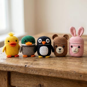 May include: Five crocheted animal-shaped containers: a yellow duck, a gray and green duck, a black and white penguin, a brown bear, and a pink bunny. Each container has a unique animal face and is sitting on a wooden surface.