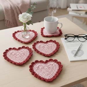 May include: Handmade crochet heart-shaped coasters in pink and burgundy. The coasters are arranged on a light-coloured wooden table, with a white mug, a vase with a white rose, and a pair of glasses. The coasters are approximately 10 cm in diameter.