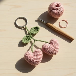 May include: A handmade crochet keychain featuring two pink heart-shaped cherries with green leaves. A silver key ring, a ball of pink yarn, a crochet hook, and a pink hair tie are also visible on a light wooden surface.