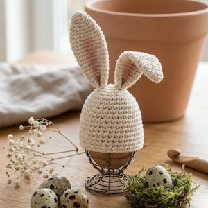 Op de afbeelding: Een crèmekleurige gehaakte eierwarmer met konijnenoren, zittend in een eierdopje van draad. De oren hebben een roze binnenkant. Een gevlekt kwartelei rust in een mosnest. Extra kwarteleieren en een terracotta pot staan op de achtergrond.