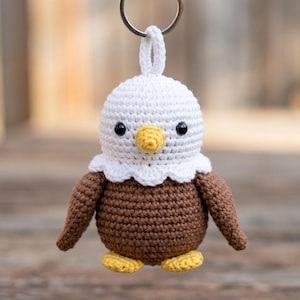 May include: A crocheted bald eagle keychain featuring a white head, a brown body, a yellow beak, and yellow feet. The eagle has black plastic eyes and a scalloped white collar detail. A silver metal keyring is attached to the top of the head.