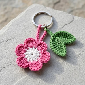 Può includere: Un portachiavi all'uncinetto con un fiore rosa dal centro bianco e una foglia verde separata. Il fiore e la foglia sono collegati a un anello in metallo color argento. Il design è composto da punti di filato nei colori rosa, bianco e verde.