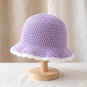 Puede incluir: Un sombrero de cubo de ganchillo lavanda con un borde festoneado blanco. El sombrero se exhibe en un soporte de sombrero de madera. El sombrero está hecho a mano y tiene un estilo clásico e informal.