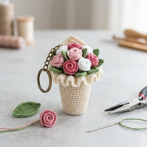 Peut inclure: Un porte-clés fait à la main en crochet représentant un bouquet de roses roses et blanches dans un pot beige avec un bord à volants. Le porte-clés comprend un anneau et une chaîne de couleur bronze. Une seule rose et une feuille au crochet sont également visibles.
