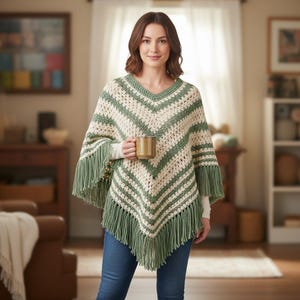 May include: A cream and green crochet poncho with a V-neck and fringe detail. The poncho has horizontal stripes and is worn by a person holding a brown mug. The style is bohemian and cozy, perfect for cooler weather.