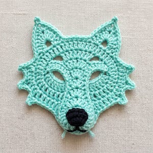 Puede incluir: Una aplicación de ganchillo en color verde menta claro con forma de cabeza de zorro o lobo. El diseño presenta orejas puntiagudas, dos aberturas para los ojos y una pequeña nariz redonda de hilo negro sólido cosida en el centro del hocico.