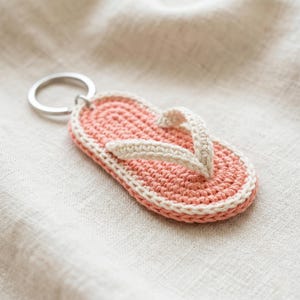 Può includere: Un portachiavi all'uncinetto fatto a mano a forma di infradito. L'infradito è principalmente rosa corallo con un bordo e un cinturino bianchi. L'anello portachiavi è argentato.