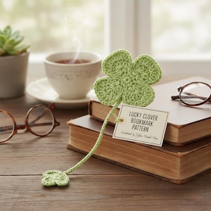 May include: A green crocheted lucky clover bookmark with a stem and a small clover at the end. The bookmark is attached to a tag that reads "LUCKY CLOVER BOOKMARK PATTERN". The bookmark rests on a stack of books.