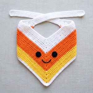May include: A triangular crochet bandana featuring a candy corn colour pattern of white, orange and yellow horizontal stripes. The accessory includes two long white ties at the top and a stitched black yarn face with two circular eyes and a smile.