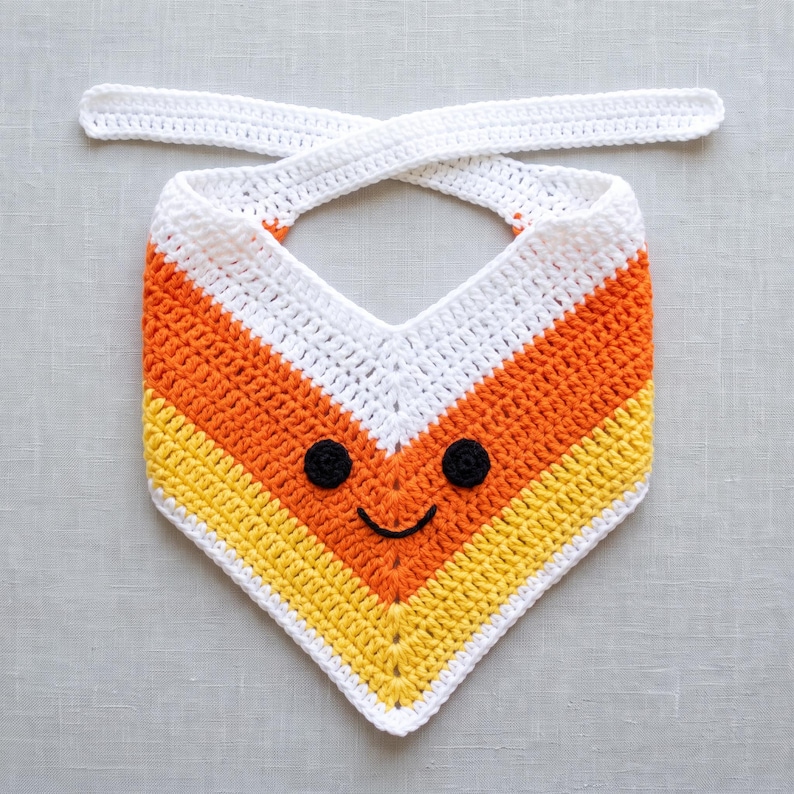 May include: A triangular crochet bandana featuring a candy corn colour pattern of white, orange and yellow horizontal stripes. The accessory includes two long white ties at the top and a stitched black yarn face with two circular eyes and a smile.