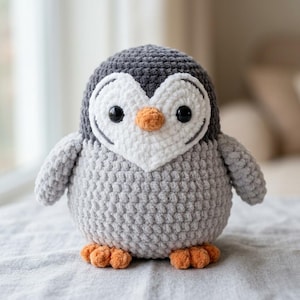 May include: A round, crocheted penguin plush toy featuring a dark grey head, a light grey body, and a white heart-shaped face patch. The penguin has two black plastic eyes, a small orange beak, and two orange feet at the base of the figure.