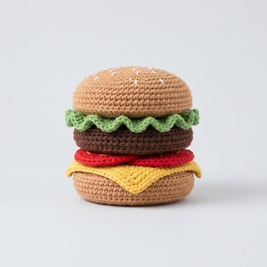 May include: A crocheted burger with a tan bun, green lettuce, a brown patty, red tomato, and yellow cheese. The burger is handmade and has white seed details on the top bun. The burger is a food-themed item.