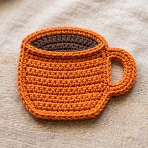 Op de afbeelding: Een platte, ronde gehaakte onderzetter in de vorm van een koffiemok. Het hoofdgedeelte van de mok is gemaakt van fel oranje garen, terwijl het binnengedeelte is gemaakt van donkerbruin garen. Het heeft een klein, afgerond handvat aan de zijkant.