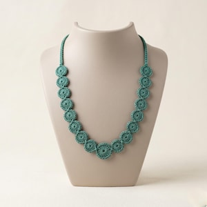 May include: A teal crochet necklace displayed on a mannequin. The necklace features a series of circular, openwork crochet elements connected by a matching cord. The necklace is a delicate piece of handmade jewelry.