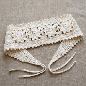 Könnte beinhalten: Ein cremefarbenes Häkelstirnband oder Gürtel mit einer zentralen Reihe aus vier detaillierten Blumenmotiven. Das Stück hat einen gewellten Rand an der Ober- und Unterseite sowie lange, dünne Häkelbänder an den Enden zum sicheren Binden.