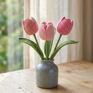 May include: A small, gray ceramic vase holds three crocheted tulips. The tulips are various shades of pink, with green stems and leaves. The vase sits on a wooden surface, with a window and curtains in the background.