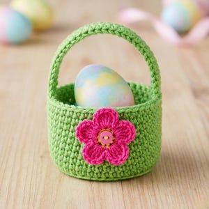 Peut inclure: Un panier de Pâques vert crocheté avec une fleur rose et un bouton. Le panier contient un œuf de Pâques aux couleurs pastel. Le panier a une anse et est posé sur une surface en bois.