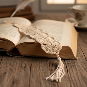 Op de afbeelding: Een crèmekleurig, gehaakt bladwijzer met een kwastje ligt op de pagina's van een open boek. De bladwijzer heeft een delicaat, open ontwerp met een geschulpte rand. Het boek ligt op een rustiek houten oppervlak, met een kop en schotel op de achtergrond.