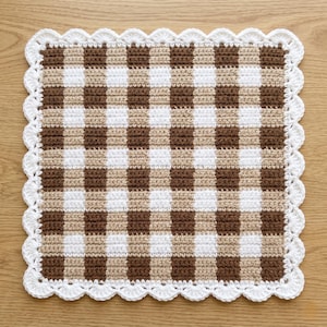 May include: A square, handmade crochet potholder with a brown, white, and tan checkered pattern. The edges are finished with a white, scalloped border. The potholder is displayed on a wooden surface.