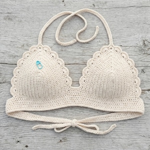 May include: Cream-colored crochet top with scalloped edges and halter straps. The handmade top features a tie closure at the back and a small blue stitch marker. The top is made of a soft, textured yarn, perfect for summer wear.