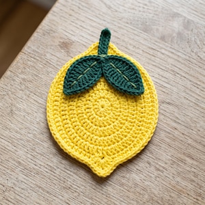May include: A lemon-shaped coaster crafted from bright yellow yarn with a circular crochet pattern. At the top, two dark green crochet leaves are attached, featuring visible light green stitched veins and a small central stem loop for hanging.