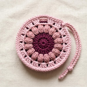 May include: A handmade, round crochet pouch in shades of pink and burgundy. The pouch features a floral design in the centre, with a drawstring closure. The pouch is approximately 10 cm in diameter.