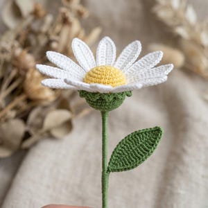 Op de afbeelding: Een handgehaakte madeliefbloem met witte bloemblaadjes, een gele kern en een groene stengel en blad. De bloem is gemaakt met een getextureerde steek, die de details van het handgemaakte item laat zien. De madelief is een charmant decoratief stuk.