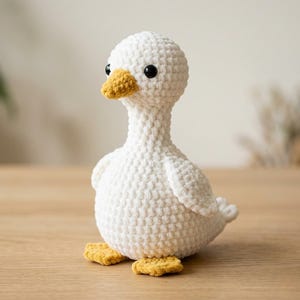 May include: A handmade crochet duck toy crafted from white yarn with a textured, looped stitch pattern. The duck features a mustard yellow beak and matching webbed feet, along with two small, round, glossy black plastic eyes set into the head.