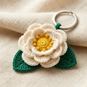 Op de afbeelding: Een handgemaakte gehaakte bloem sleutelhanger. De bloem heeft crèmekleurige bloemblaadjes met een gele kern en groene bladeren. De sleutelhanger is bevestigd aan een zilveren metalen ring.