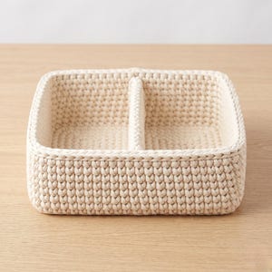 May include: A cream-colored, rectangular crochet basket with a central divider, creating two compartments. The basket is made of thick, textured yarn and sits on a light wooden surface. The design is simple and functional, suitable for organizing small items.