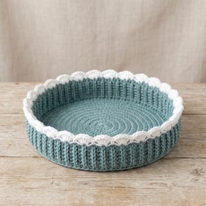 May include: A round, teal-colored crochet basket with a white scalloped edge. The basket is made of thick yarn and sits on a wooden surface. The interior of the basket has a spiral pattern.