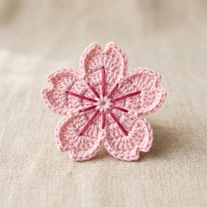 May include: A handmade, light pink crochet flower with five petals. The center of the flower features dark pink embroidered details. The flower is set against a neutral, textured background, highlighting the delicate craftsmanship.