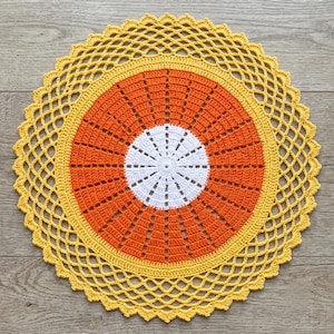 May include: A round, handmade crochet doily featuring a central white circular section, a wide middle band of vibrant orange yarn, and an outer border of bright yellow yarn with an intricate, open-work lattice pattern and a scalloped edge.