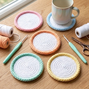 Op de afbeelding: Vijf ronde gehaakte onderzetters met witte centra en kleurrijke gevlochten randen in roze, blauw, perzik, mintgroen en geel. Een keramische mok staat op de blauwe onderzetter. Twee haaknaalden, twee klossen garen en een metalen schaar zijn aanwezig.
