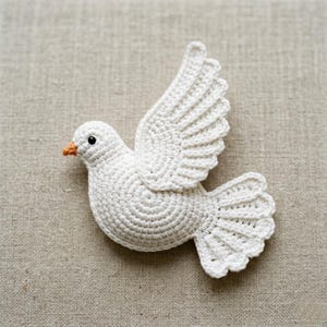 Könnte beinhalten: Eine gehäkelte weiße Taube mit einem kleinen orangefarbenen Schnabel und einem einzelnen schwarzen Perlenauge. Der Vogel ist im Seitenprofil dargestellt, mit erhobenem Flügel und gefächerten Schwanzfedern, die detaillierte Garnstiche zeigen.