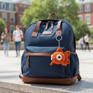 May include: A navy blue backpack with brown leather accents and a crocheted orange goldfish keychain. The backpack has multiple pockets and a top handle. The keychain is attached to the zipper.
