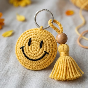 May include: A yellow crocheted smiley face keychain with a black stitched mouth and eyes. The keychain has a silver ring, a wooden bead, and a yellow tassel. The background is a neutral fabric.