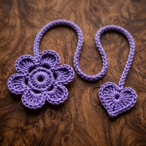 May include: Two handmade crochet items: a flower and a heart, both in a vibrant purple hue. The flower has multiple petals, while the heart is a simple, rounded shape. Both are connected to a braided cord, suggesting they could be used as bookmarks or decorative accents.