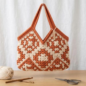 Może przedstawiać: Ręcznie robiona torba typu tote wykonana na szydełku z wzorem granny square. Torba ma geometryczny wzór w kolorach rdzawo-pomarańczowym i kremowym, z pasującymi uchwytami. Do uchwytu przymocowana jest mała metka. Widoczne są szydełka, przędza i nożyczki.