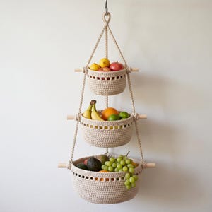 May include: A three-tiered hanging fruit basket made of beige crochet material. Each basket is supported by wooden dowels and suspended by rope. The baskets are filled with various fruits, including apples, bananas, oranges, and grapes.
