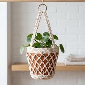 May include: A cream-colored, crocheted plant hanger with a diamond pattern, holding a terracotta pot filled with green leafy plants. The hanger is suspended from a brass ring and a wooden rod, creating a stylish display for indoor plants.