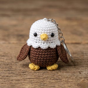 May include: A crocheted eagle keychain with a white head, brown body and wings, and yellow beak and feet. The keychain has a silver metal ring and chain. The eagle is standing on a wooden surface.