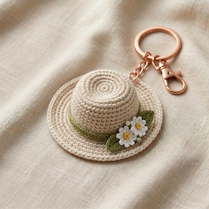 Puede incluir: Un llavero de sombrero de ganchillo beige con una banda verde y dos pequeños acentos de margaritas blancas. El llavero tiene un cierre y un anillo de color cobre. El sombrero mide aproximadamente 5 cm de diámetro.