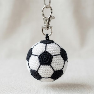 Könnte beinhalten: Gehäkelter Fußball-Schlüsselanhänger. Der Ball ist weiß mit schwarzen Fünf- und Sechsecken und an einem silberfarbenen Metallverschluss befestigt. Ein niedliches Accessoire für Sportfans.