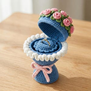 May include: A handmade blue crochet ring box with a white scalloped trim and a pink bow. The lid is decorated with pink and green crochet flowers, and it holds a gold ring.