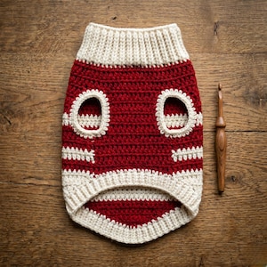 Puede incluir: Un suéter para perro de ganchillo hecho a mano, rojo y crema, con agujeros para los brazos. El suéter tiene un cuello y un dobladillo inferior de color crema acanalado. Un ganchillo de madera descansa sobre la superficie de madera junto al suéter.