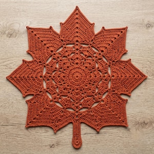 Puede incluir: Un tapete de ganchillo color óxido con forma de hoja de arce grande. El intrincado diseño presenta un patrón central circular tipo mandala que se transforma en lóbulos de hoja puntiagudos y radiantes con calados en toda la pieza.