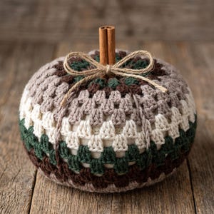 May include: A round, stuffed pumpkin decoration featuring a crocheted granny square cover in alternating bands of dark brown, forest green, cream, and taupe yarn. A cinnamon stick serves as the stem, tied with a thin, natural jute twine bow.
